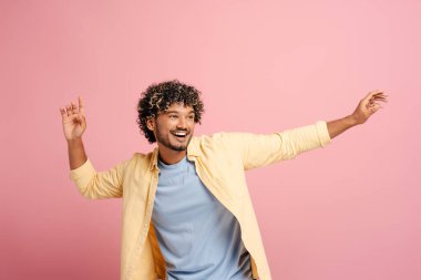 Kıvırcık saçlı genç adam pembe bir arka planda mutlu bir şekilde dans eder, neşe ve kaygısızlıkla el kol gezer.