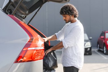 Genç, Hintli bir adam arabasını topluyor ve yolculuk için hazırlanıyor. Bavullarını bir otoparktaki bagaja koyuyor.