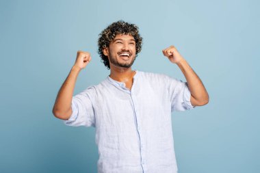 Kıvırcık saçlı genç adam kollarını havaya kaldırıyor ve gülümsüyor, başarıyı ve başarıyı neşeli bir ifadeyle kutluyor.