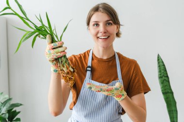 Önlüklü ve eldivenli neşeli bahçıvan kadın gülümser, köklerini göstermek için bir bitki tutar, özgüven ve pozitiflikle bakım ipuçlarını paylaşır.