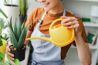 Kadın çiçekleri sarı bir sulama tenekesiyle sulayarak onlara bakıyor. Gülümsüyor çünkü bitkilerle ilgilenmeyi seviyor.