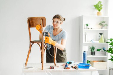 Kadın marangoz, konforlu atölyesinde eski bir sandalyeyi restore etmek için elektrikli zımpara makinesi kullanıyor.