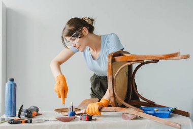 Sanatçı kadın, atölyesindeki aletler ve malzemelerle çevrili eski bir ahşap sandalyeyi restore etmek için bir keski kullanıyor. Koruyucu gözlük ve eldiven takıyor.