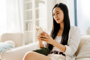 Gülümseyen, güzel Asyalı kadın oturma odasında bir kanepede oturuyor, akıllı telefonunu kullanıyor. Çevrimiçi alışveriş kavramı, sosyal medya