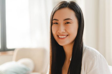 Güzel gülümseyen Asyalı kadın oturma odasında dinleniyor, kameraya bakıyor, yakınlaşıyor. Reklam kavramı