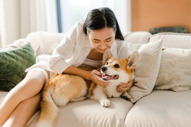 Asyalı, gülümseyen, mutlu bir kadın kanepede dinleniyor ve mutlu Corgi köpeğini evde okşuyor. Hayvan sevgisi konsepti