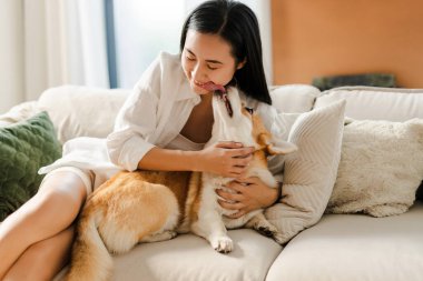 Çekici, gülümseyen bir kadın kanepede dinleniyor ve mutlu Corgi Pembroke köpeğini kucaklıyor. Hayvan sevgisi konsepti