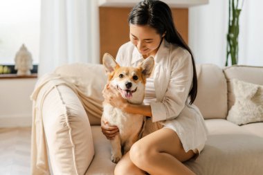 Genç Asyalı bir kadın modern dairesinde rahat bir kanepede oturuyor, tatlı Corgi köpeğine sarılırken rahatlatıcı bir anın tadını çıkarıyor.