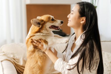 Gülümseyen Asyalı kadın rahat bir kanepede oturuyor, sevimli Corgi köpeğini tutuyor, sevgi ve neşeyi paylaşıyor. Görüntü, evcil hayvan ve sahibi arasındaki bağı sıcak bir ortamda yakalıyor.