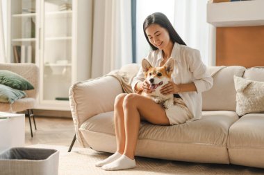 Genç, gülümseyen Asyalı kadın kanepede dinleniyor ve evcil köpeğiyle kucaklaşıyor, birlikte vakit geçiriyor. Hayvan konseptine bayılırım.
