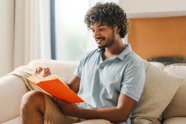Mutlu, gülümseyen kıvırcık saçlı Hintli adam evdeki kanepede kitabın tadını çıkarıyor, sayfayı çeviriyor. Hobi konsepti
