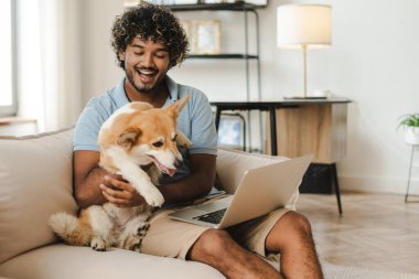 Neşeli köpeğini tutan gülümseyen Hintli genç adam dizüstü bilgisayarla evde çalışıyor, video izliyor, birlikte vakit geçiriyor. Aşk hayvanı, online teknoloji konsepti.