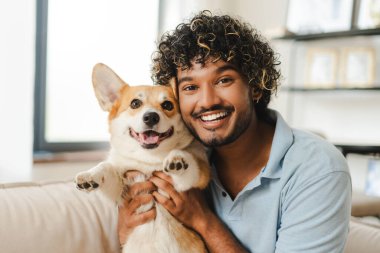 Evdeki kanepede sevimli Corgi köpeğini tutarken mutlu bir şekilde gülümseyen bir adam, doğal ışıkla dolu rahat bir oturma odasında sevgi ve dostluk yayıyor.