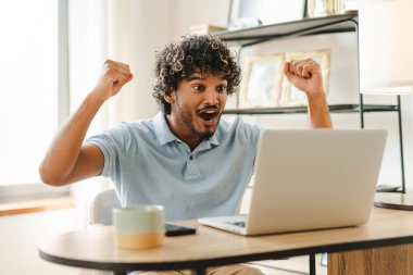 Young Indian man celebrates his success while working from home, surrounded by technology and a cup of coffee, in a cozy room filled with natural light clipart