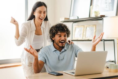 Heyecanlı çok ırklı çift, evde dizüstü bilgisayar başında çalışırken modern uzak yaşam tarzlarını sergileyerek başarıyı kutluyor. Aşkın ve birlikteliğin göstergesi.