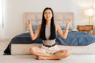 Genç bir kadın yatak odasında yerde oturuyor, bacakları çapraz ve gözleri kapalı, stresi azaltmak ve akıl sağlığını iyileştirmek için meditasyon yapıyor. Uyum kavramı