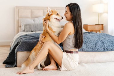 Genç kadın, yatak odasının rahatlığında sevimli corgi köpeğiyle hassas bir anı paylaşıyor. Barışçıl ve sevgi dolu bir ortamda birbirlerine eşlik etmekten zevk alıyorlar.