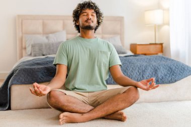 Genç Hintli adam nilüfer pozisyonunda yerde oturuyor, yatak odasında gözleri kapalı meditasyon yapıyor. Farkındalık yapıyor ve huzurlu görünüyor.