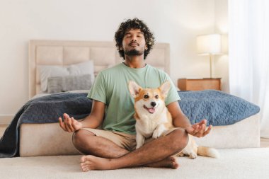 Genç, Hintli bir adam Lotus Yoga pozisyonunda gözleri kapalı otururken köpeği yatak odasında kucağında oturuyor.