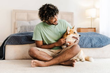 Genç, gülümseyen Hintli adam, Corgi 'sini sıcak bir yatak odasında mutlu bir şekilde okşuyor. Sevgi ve neşe saçan bir arkadaşlık ve rahatlama anı.