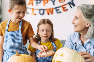 İki neşeli çocuk önlük giyip balkabağı çekirdeğini kepçelerken büyükanneleriyle evde balkabağı kabağı oyuyorlar. Cadılar bayramını kutlayıp eğleniyorlar.