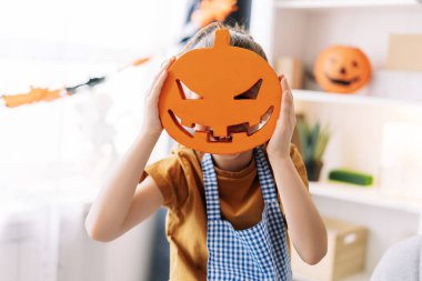 Yüzlerinin önünde oyulmuş balkabağı süslemesi tutan çocuk önlük takıyor. Arka plan cadılar bayramı için dekore edildi. Kutlama konsepti