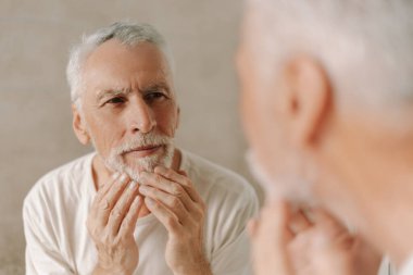 Beyaz saçlı ve sakallı yaşlı bir adam aynaya bakıyor ve sakalına dokunuyor, yaşını ve görünüşünü yansıtıyor. Cilt bakımı kavramı