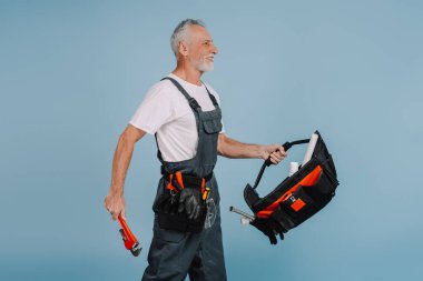 Boru anahtarı ve alet çantası taşıyan yetişkin bir tesisatçı işe yarayacak. Alet kemeri takıyor ve kamera dışında bir şeye gülümsüyor.