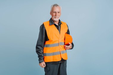 Daha yaşlı bir inşaat işçisi yüksek görüş mesafesi olan bir yelek giyiyor. Kaskını tutuyor ve mavi arka planda kameraya gülümsüyor.