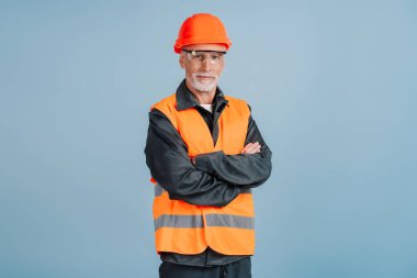 Başlık ve güvenlik yeleği giyen deneyimli inşaat işçisi kollarını kavuşturarak mavi arka planda güven ve uzmanlık aktarıyor.