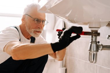 Lavabo borusunu tamir eden, eldiven takan ve güvenlik gözlüğü takan yetenekli bir tesisatçı. Sızdıran banyo borularını özenle onarmak, hassasiyet ve uzmanlık gösterisi.