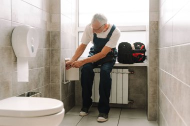 Mavi tulumlu yetenekli tesisatçı modern bir banyoda tuvalet depolarında çalışıyor. Tesisatı tamir etmek için tornavida kullanıyor.