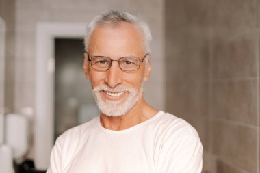 Beyaz sakallı, mutlu, sağlıklı, olgun bir adam banyo ortamında gülümsüyor. Beyaz bir gömlek ve gözlük takıyor.