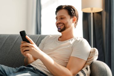 Genç adam mutlu bir şekilde akıllı telefonunu evdeki rahat bir kanepede kullanıyor. Kaygısız bir günün tadını çıkarıyor.