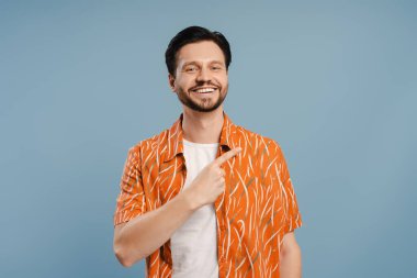 Yakışıklı genç sakallı adam kopya alanını işaret ediyor ve mavi arka planda izole bir şekilde gülümsüyor. Reklam kavramı
