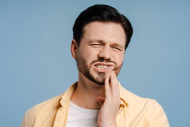 Güçlü diş ağrısı çeken üzgün bir adam mavi arka planda yanağa dokunuyor, tedavi oluyor. Diş sağlığı konsepti