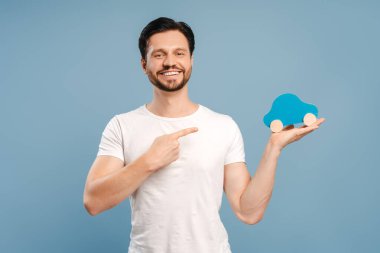 Heyecanlı adam mavi oyuncak arabasıyla beyaz stüdyoyu işaret ediyor. Araba reklamları, pazarlama ya da promosyonlar için mükemmel. Mavi tişört uyuyor, çok yönlü tasarım.