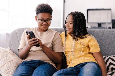 İki arkadaş evde akıllı bir telefondan sosyal medyada geziniyor, oturma odasında kaynaşıyorlar. Afro-amerikan gençler birlikte gündelik eğleniyorlar.