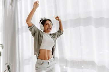 Genç kadın evde eğleniyor, kulaklık takıyor, kolları havada ve gözleri kapalı dans ediyor.