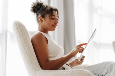 Genç bir kadın rahat bir sandalyede oturuyor, gülümsüyor ve dijital tablet kullanıyor.