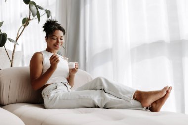 Genç kadın evdeki kanepesinde dinleniyor, bir fincan yoğurdun tadını çıkarıyor ve gülümsüyor.