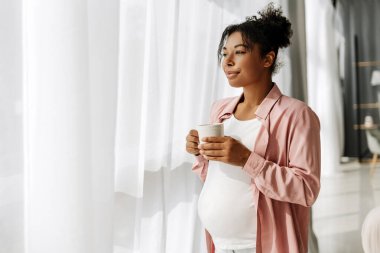 Elinde kupa tutan hamile bir kadın yatak odasında dikilirken düşünceli bir şekilde pencereden bakıyor.