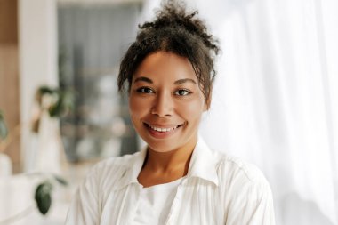 Genç siyah kadın rahat evinde mutlu bir şekilde gülümsüyor, kendine güveni tam ve tasasız bir tavırla. Kahverengi gözleri ve kıvırcık saçları var, günlük beyaz bir gömlek giyiyor, doğrudan kameraya bakıyor.