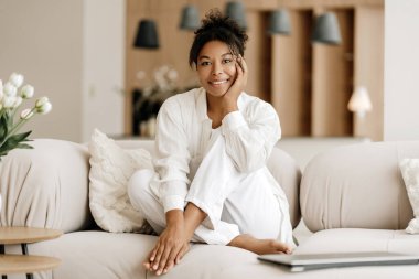 Genç bir kadın, eli çenesinde rahat bir kanepede oturuyor, gülümsüyor ve dizüstü bilgisayarı yanında.