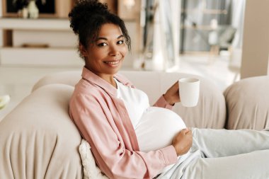 Hamile kadın rahat bir koltukta dinleniyor, evinde sıcak bir fincan çayın tadını çıkarıyor.