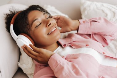 Genç bir kadın, gözleri kapalı bir kanepede uzanıyor kulaklıklarıyla müziğin tadını çıkarıyor.