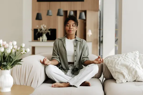Genç kadın, modern oturma odasındaki rahat bir kanepede meditasyon yaparken farkındalık ve huzurun tadını çıkarıyor.