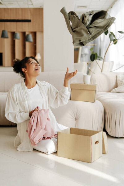 Cheerful black woman throwing clothes in donation box, feeling happy and grateful while decluttering wardrobe at home