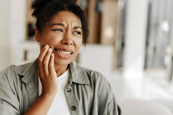 Genç bir kadın yanağına dokunuyor ve güçlü bir diş ağrısı hissediyor oturma odasında bir kanepede oturuyor.