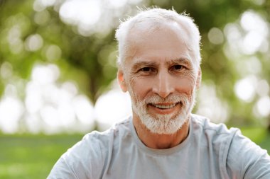 Parkta güneşli bir günün tadını çıkarırken gülümseyen mutlu, kır saçlı adamın portresi, kamera fotokopi alanına bakıyor. Reklam kavramı